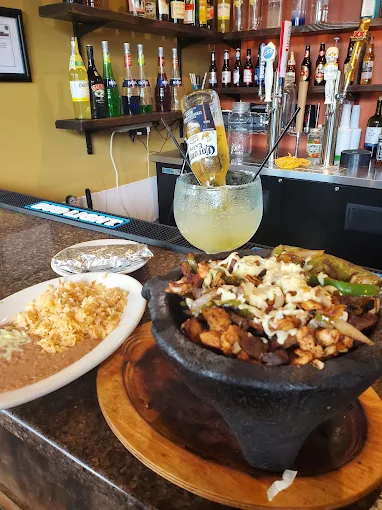 A stone bowl of fajita-style meat and vegetables sits on a bar next to a plate of rice and beans, accompanied by a margari...